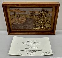Frontal photo of metal relief sculpture framed and standing on a white surface with certificate of authenticity and artist's letter beneath it.