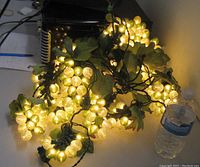 Full view of glowing grape bunch lights with leaves on a table beside a water bottle for scale showing multiple bunches lit in warm white.