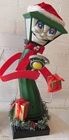 Front side view of animated lamp post figurine "Frankie" wearing Santa hat and scarf, holding a wrapped gift box. Shows details of facial expression and decorations on base.