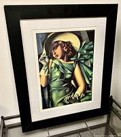 Framed art print showing an Art Deco style portrait of a woman in green dress and hat with gloves, in a black frame with off-white mat.