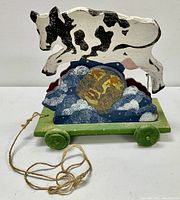 Front view of wooden pull toy with cow figure mounted above an illustrated moon with clouds on wheeled base, showing attached pull string.