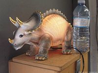 Side view of the ceramic triceratops lamp illuminated, showing detailed textures, golden highlights on horns, toes and frill, next to a water bottle for size comparison.