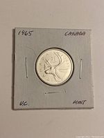 Reverse side of the 1965 Canadian silver quarter showing the caribou design with text 'CANADA 25 CENTS 1965' in a white protective cardboard holder.