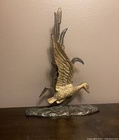 Front angled view of brass and bronze duck figurine mounted on base with reeds and water detail.