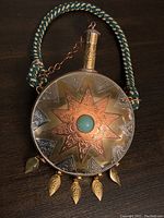 Front view of copper flask showing detailed star pattern, central turquoise cabochon, and leaf charms hanging below. Braided rope handle shown.