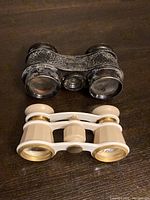 Two pairs of vintage opera glasses on wooden surface; one black textured and worn, the other cream-colored with gold accents.