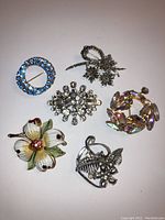 Photo shows six rhinestone brooches arranged on a white background, highlighting their design and condition
