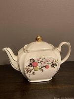 Side view of white china teapot with embossed floral apple blossom and apple decoration and gold finial knob on lid