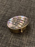 Photo showing top view of the small Mexican oval box with iridescent shell inlay lid and metal body on textured surface.