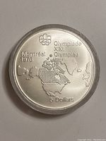 Front side of the 1976 Montreal Olympic silver coin showing the Olympic rings, map of Canada, and denomination, enclosed in a clear protective case.