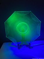 Turquoise octagonal uranium vaseline glass plate glowing bright green under black light on a display stand, showing Art Deco etched pattern detail.