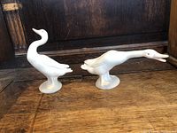 Side view of two white ceramic Lladro swan figurines on wood surface, showing distinct neck postures and detailing.