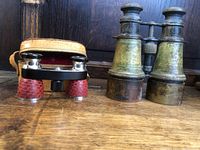 Two pairs of binoculars set side by side, showing the small opera glass binocular with red grips and case, and the larger brass binocular with patina.
