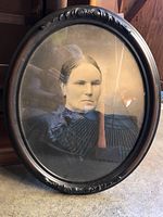 Front view of the oval framed black and white portrait photo of a woman, showing the decorative black carved frame and the woman's facial expression and attire.