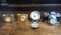 All four items displayed in a row on a wooden surface: three camera-shaped clocks and one travel watch with National Geographic logo.