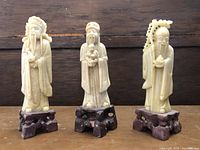 Three ivory-toned Chinese soapstone carved figures on wooden bases, showing overall condition and style.