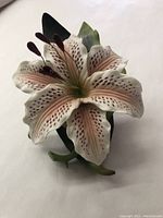 Side angle view of the ceramic lily flower sculpture showing delicate pink and white petals with dark spots and stamens, and surrounding green leaves.