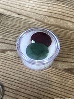Photo of loose ruby and emerald gemstones inside small clear container on wooden surface.