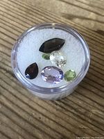 Top view of various loose gemstones on white foam in round clear plastic container showing assortment of colors and shapes