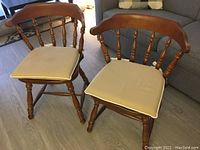 Two vintage Windsor style wooden chairs with tan cushions sitting on wood laminate floor near a sofa and window.