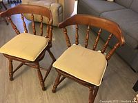 Front view of two Windsor style wooden chairs with beige cushions, showing spindle backs and turned legs.