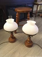 Photo showing two vintage hurricane lamps with ribbed white glass shades and wooden bases, in front of a wooden stool.