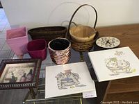 Wide view of all lot contents on a glass table including baskets, vases, and wall art (plates and framed shadow box)