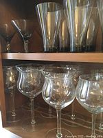 Photo of crystal and glassware on a wooden shelf showing tall cylindrical tumblers, stemmed glasses with broad bowls and some cocktail glasses.