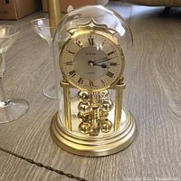 Front view of German dome clock with visible face, Roman numerals, gold base, and rotating pendulum under a glass dome.
