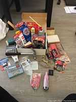 Top view showing assorted tools, batteries, hardware and small accessories arranged on floor, including a claw hammer, brass handles, new batteries in original packaging, hand tools and consumables.