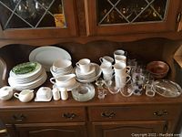 Overall view of porcelain dishware set including plates, mugs, bowls, creamer, sugar bowl, butter dish, and assorted glassware including decorated glasses and stemmed glasses, displayed on wooden cabinet.