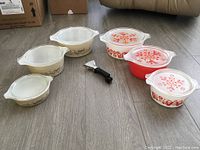 Overview of two vintage Pyrex casserole sets with lids and black lid holder on floor.