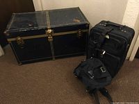 Black steamers trunk alongside two pieces of luggage on carpeted floor.