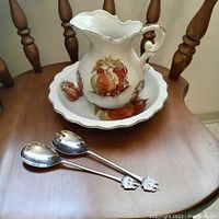 Photo showing vintage ceramic wash basin and pitcher with fruit motif alongside two long-handled spoons with crest emblems