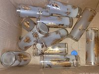 Box containing 11 tall, slender frosted glass juice cups with gold leaf patterns and gold trim around the rim, arranged in various positions inside the box.