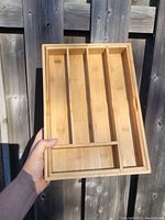 Photo showing bamboo cutlery tray with four compartments for utensils, held horizontally in hand against wooden fence background.