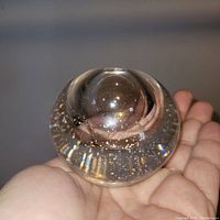 Photo of clear round blown glass paperweight with natural gold flakes held in hand, showing top and side view with dimple base.