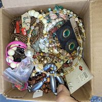 Top-down view of a box filled with assorted costume jewelry showing various necklaces, bracelets, earrings, and small jewelry components in mixed materials and colors.