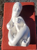 Photo of the full ceramic bust showing two lovers embracing in white matte ceramic on an orange cloth background outdoors.