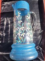 Full view of hand painted blown glass pitcher showing blue rim and base with white floral decoration on clear glass body.