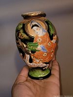 Front and angled view showing hand painted depiction of a Japanese figure in traditional dress with gold trim on the porcelain vase held in hand for size reference.
