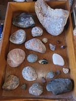 Top down view showing all mineral and rock specimens in a wooden tray, highlighting a large rough textured specimen and smaller rounded specimens with varied colors and textures.