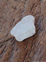Close up image of a white translucent natural sea glass piece about 1 inch with textured frosted surface and irregular shape, placed on textured brown surface.