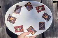 Oval white ceramic serving platter with eight vintage label-style designs arranged around inner rim. Labels include 'SOLABELLA', 'FLORIO CINZANO', 'BARONI' with illustrations of pin-up figures.