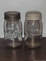 Two antique GEM Mason jars, one light grey clear and one aqua clear, both with metal lids, standing side by side on a light surface.