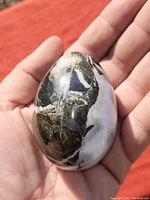 Hand holding a polished stone egg with visible black, white, and greenish natural swirl patterns under sunlight showing the reflective glossy finish.