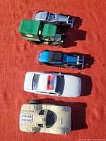 Five vintage toy cars arranged on orange surface, showing top and side views, colors include green, silver, blue, white, and tan.