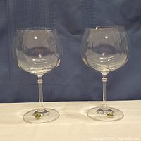 Pair of 2 clear Bohemia crystal wine glasses on white surface with blue background. Both glasses have gold and black Bohemia Crystal stickers on their bases.