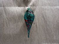 Single elongated teardrop-shaped art glass hanging ornament, swirled green, red, and black colors, clear hanging loop top, on a textured gray fabric background.