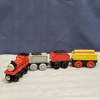 Four connected wooden toy train pieces showing from the side on light surface, depicting James the Red Engine and cars